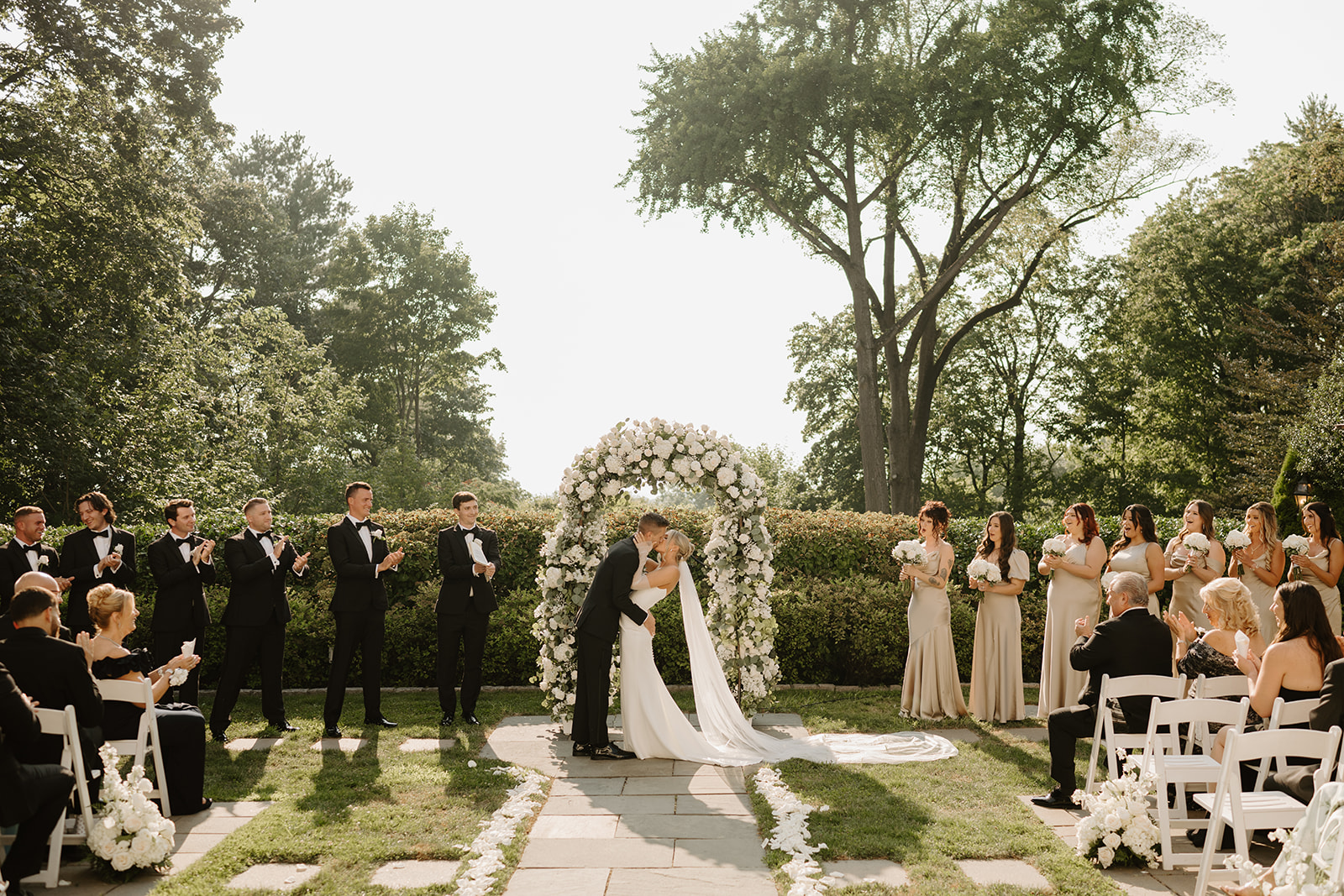Bride and groom share their first kiss under a white rose arch, as the wedding party applauds.